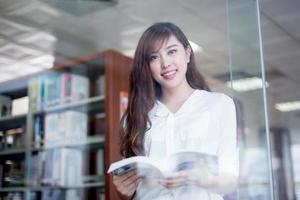 Estudiante asiática hermosa que sostiene el libro en retrato de la biblioteca foto