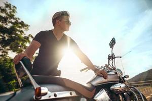 Motorcyclist stands next to bike, backlit by sun photo