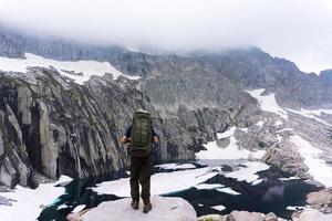 Hombre con mochila en acantilado frente al agua y la montaña