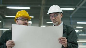 två ingenjörer i hårdhattar diskuterar en ritning när de står i en fabrik.