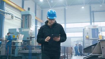 Factory worker in a hard hat is walking and using a tablet computer.