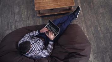 homem deitado em uma poltrona, segurando um laptop e digitando