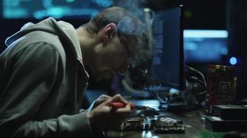 elektronica-ingenieur soldeert een elektrisch bord met processors in een donker kantoor met beeldschermen.