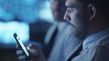 säkerhetsansvarig kontrollerar sociala medier på smarttelefonen medan han arbetar i ett mörkt övervakningsrum fyllt med skärmar.