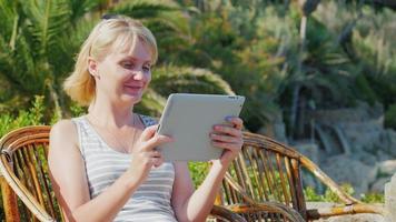 donna turista con il tablet dice. si siede su una sedia di vimini sullo sfondo delle palme. sempre connesso in un resort tropicale