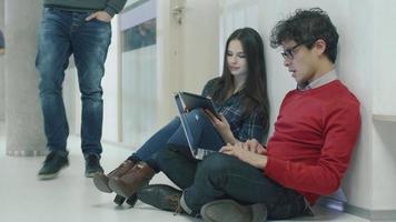Male and female students are sitting in a college hallway and working on a laptop computer and tablet.