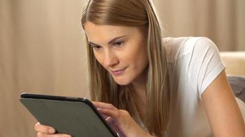 bela jovem atraente em uma camiseta branca com um computador tablet deitado em um sofá. navegando na internet e sorrindo
