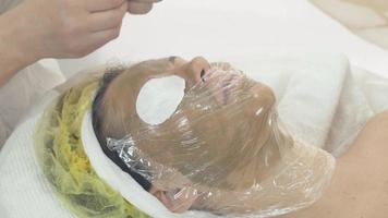 Cosmetologist put thermofilm on woman face with green mask in saloon. Cotton pad