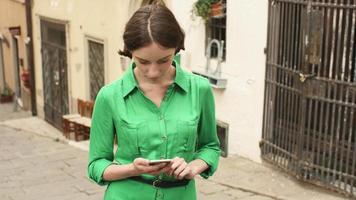 attraktiv ung brunettkvinna i ljus sommarklänning på gatorna i den europeiska staden. hon tittar på sin mobiltelefon och använder den. video