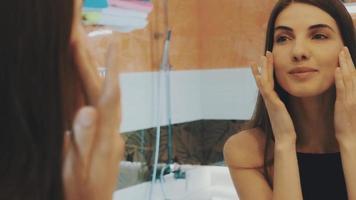 Young brunette girl looking in mirror in bathroom. Touch face, neck. Smile