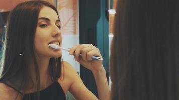 Young brunette girl brush teeth in front of mirror in bathroom. Morning hygiene