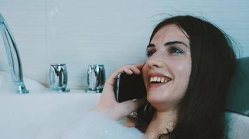 fille brune prenant un bain plein de mousse. parler au téléphone. se détendre. repos