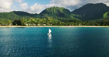 Luftaufnahme, die über traditionelles hawaiianisches Segelboot in tropischer blauer Lagune in Richtung schöner grüner Berge fliegt