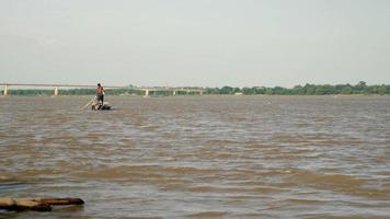 homem remando seu pequeno barco no rio enquanto puxa uma rede de pesca na água atrás