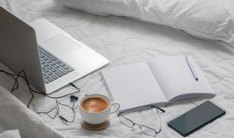 Laptop with a coffee, phone and notebook on a bed photo