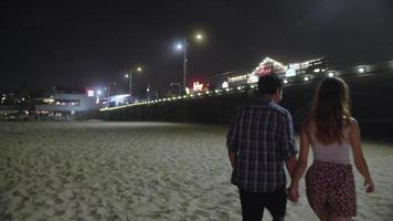 Cámara lenta de la joven pareja cogidos de la mano caminando en la playa por la noche
