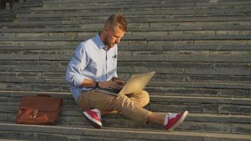 Handsome student sitting on stone steps and using a laptop