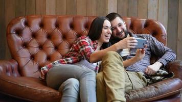 Sonriente pareja joven sentada en el sofá y tomando una foto selfie con el teléfono inteligente