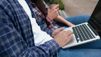 jovens urbanos em camisas xadrez e jeans azul, usando computador laptop e tomando café em uma xícara de café no banco