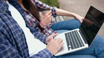 Young urban people  in checkered shirts and blue jeans using computer laptop and drinking coffee from to go cup on bench