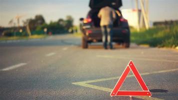 Triángulo de advertencia rojo en la carretera, avería del coche, conductor buscando herramientas en el maletero