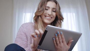 fröhliche junge Frau mit langen Haaren, die auf Bett sitzt und Tablet-Computer benutzt.
