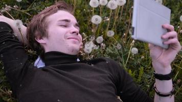 junger glücklicher Student, der Tablette im Park benutzt, im Löwenzahn auf dem Gras liegend, lächelnd und lachend