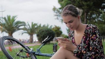 garota muito fofa usando smartphone ao lado de sua bicicleta no parque com as palmas das mãos em um dia ensolarado video