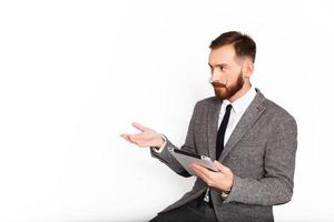 Hombre serio en traje gris gesticulando mientras sostiene una tableta foto