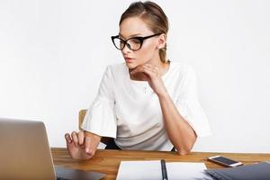Mujer pensativa trabaja en un portátil en un escritorio sobre fondo blanco.