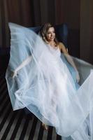 Thoughtful bride sits in a deep blue chair in dark hotel room photo