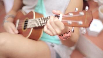 Sitting girl in shorts playing ukulele guitar on street. Summer sunny day. Music. Strings. Sound video