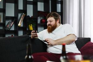 Fat man types the number of a credit card in his phone sitting on the couch photo