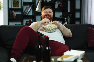 Laughing fat man sits on the sofa eating popcorn and watching TV photo