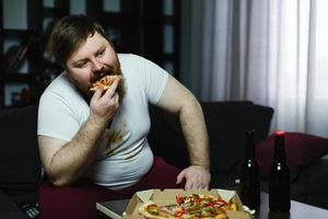 Ugly fat man eats pizza sitting on the sofa photo