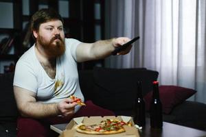 Fat man eats pizza sitting on the sofa photo