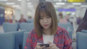 Happy Asian woman using and checking her smartphone while sitting on chair in terminal hall while waiting her flight at the departure gate in international airport. video