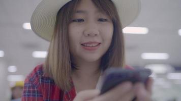 Happy Asian woman using and checking her smartphone while sitting on chair in terminal hall while waiting her flight at the departure gate in international airport. video