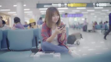 gelukkige aziatische vrouw die haar smartphone gebruikt en controleert terwijl zij op stoel in terminalzaal zit terwijl zij haar vlucht wacht bij de vertrekgate op de internationale luchthaven.