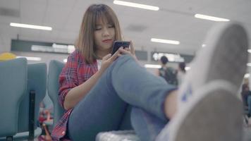 Happy Asian woman using and checking her smartphone while sitting on chair in terminal hall while waiting her flight at the departure gate in international airport. video