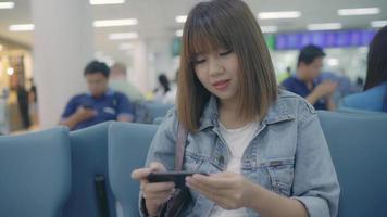 Feliz mujer asiática usando y revisando su teléfono inteligente mientras está sentada en una silla en el pasillo de la terminal mientras espera su vuelo en la puerta de salida del aeropuerto internacional. video