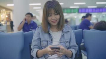 cámara lenta - feliz mujer asiática usando y comprobando su teléfono inteligente en el pasillo de la terminal mientras espera su vuelo en la puerta de salida del aeropuerto internacional. video