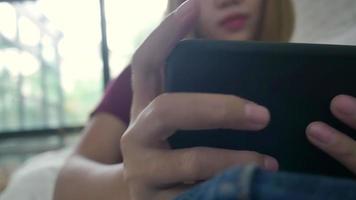 mujer asiática jugando smartphone mientras está acostado en el sofá de su casa en su sala de estar. mujer feliz usa el teléfono para enviar mensajes de texto, leer, enviar mensajes y comprar en línea en casa. video