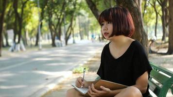 glückliche junge asiatische Frau des Hipsters, die in ihr Tagebuch im Park schreibt. video