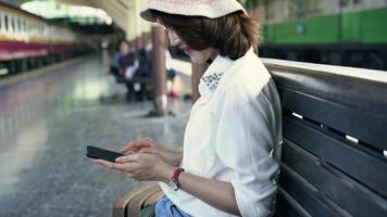 viajero, mochilero, mujer asiática, viaje, en, bangkok, tailandia. feliz mujer joven sentada en un banco con smartphone para hablar, leer y enviar mensajes de texto en la estación de tren. video