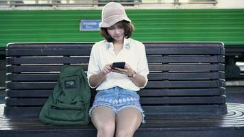 viajero, mochilero, mujer asiática, viaje, en, bangkok, tailandia. feliz mujer joven sentada en un banco con smartphone para hablar, leer y enviar mensajes de texto en la estación de tren.