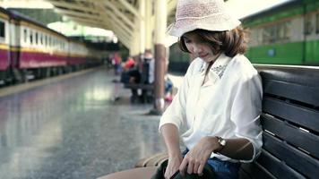 Happy teen Asian woman listening to the music with headphones and waiting in a train station summer. Travel Thailand concept. video