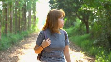 glad ung asiatisk kvinna resenär med ryggsäck promenader i skogen. vandrare asiatisk kvinna med ryggsäck som går på stig i sommarskog. äventyr backpacker resor människor koncept.
