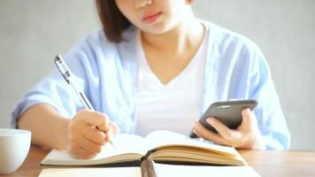 mujer de negocios joven en vestido blanco sentado a la mesa en la cafetería y escribiendo en el cuaderno. mujer asiática hablando teléfono inteligente y taza de café. autónomo que trabaja en cafetería. aprendizaje de los estudiantes en línea. video