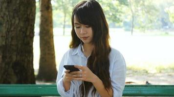 Mulher bonita está lendo mensagem de texto agradável no celular enquanto está sentada no parque em um dia quente de primavera video